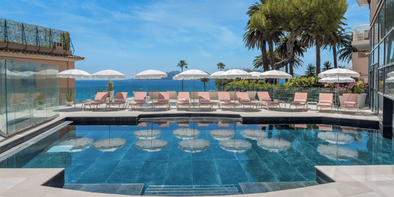 Rooftop pool area with pink lounge chairs and white umbrellas facing the blue sea and distant islands, palm trees on the edge.