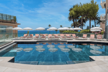 Rooftop pool area with pink lounge chairs and white umbrellas facing the blue sea and distant islands, palm trees on the edge.