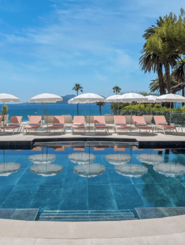Rooftop pool area with pink lounge chairs and white umbrellas facing the blue sea and distant islands, palm trees on the edge.