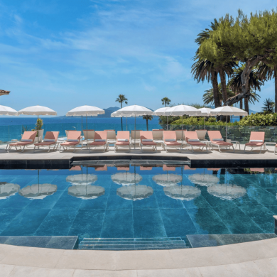 Rooftop pool area with pink lounge chairs and white umbrellas facing the blue sea and distant islands, palm trees on the edge.