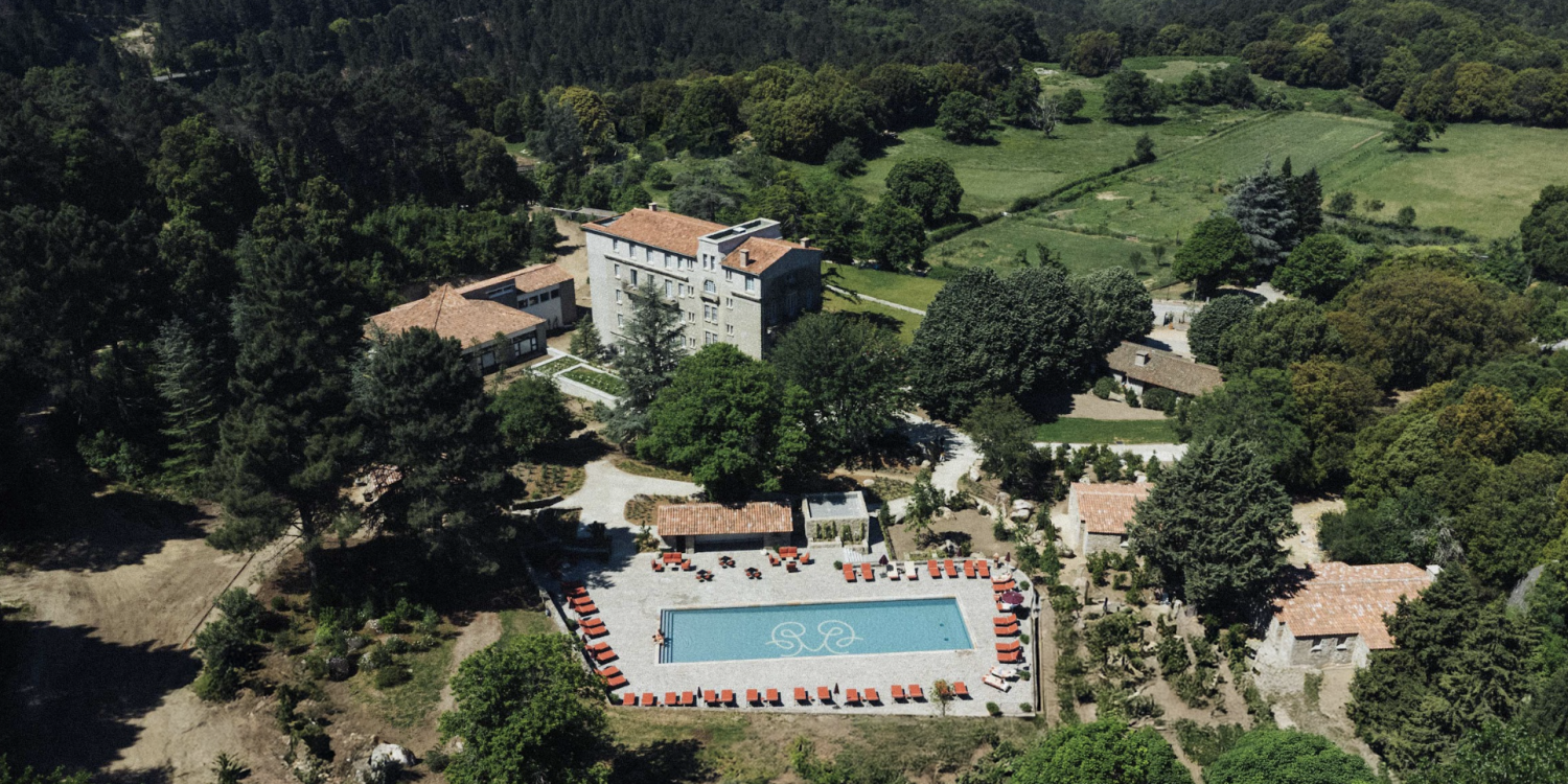 Aerial view of a resort complex: a rectangular blue swimming pool with red lounge chairs around it, stone buildings with terracotta roofs, and surrounding trees and green fields.