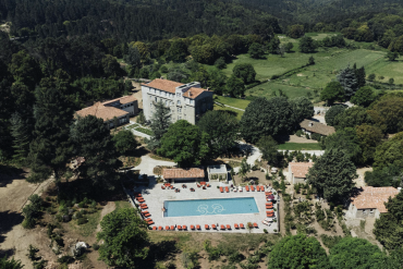 Aerial view of a resort complex: a rectangular blue swimming pool with red lounge chairs around it, stone buildings with terracotta roofs, and surrounding trees and green fields.