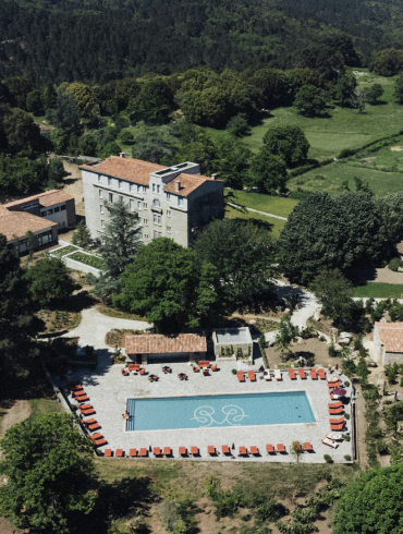 Aerial view of a resort complex: a rectangular blue swimming pool with red lounge chairs around it, stone buildings with terracotta roofs, and surrounding trees and green fields.