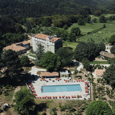 Aerial view of a resort complex: a rectangular blue swimming pool with red lounge chairs around it, stone buildings with terracotta roofs, and surrounding trees and green fields.