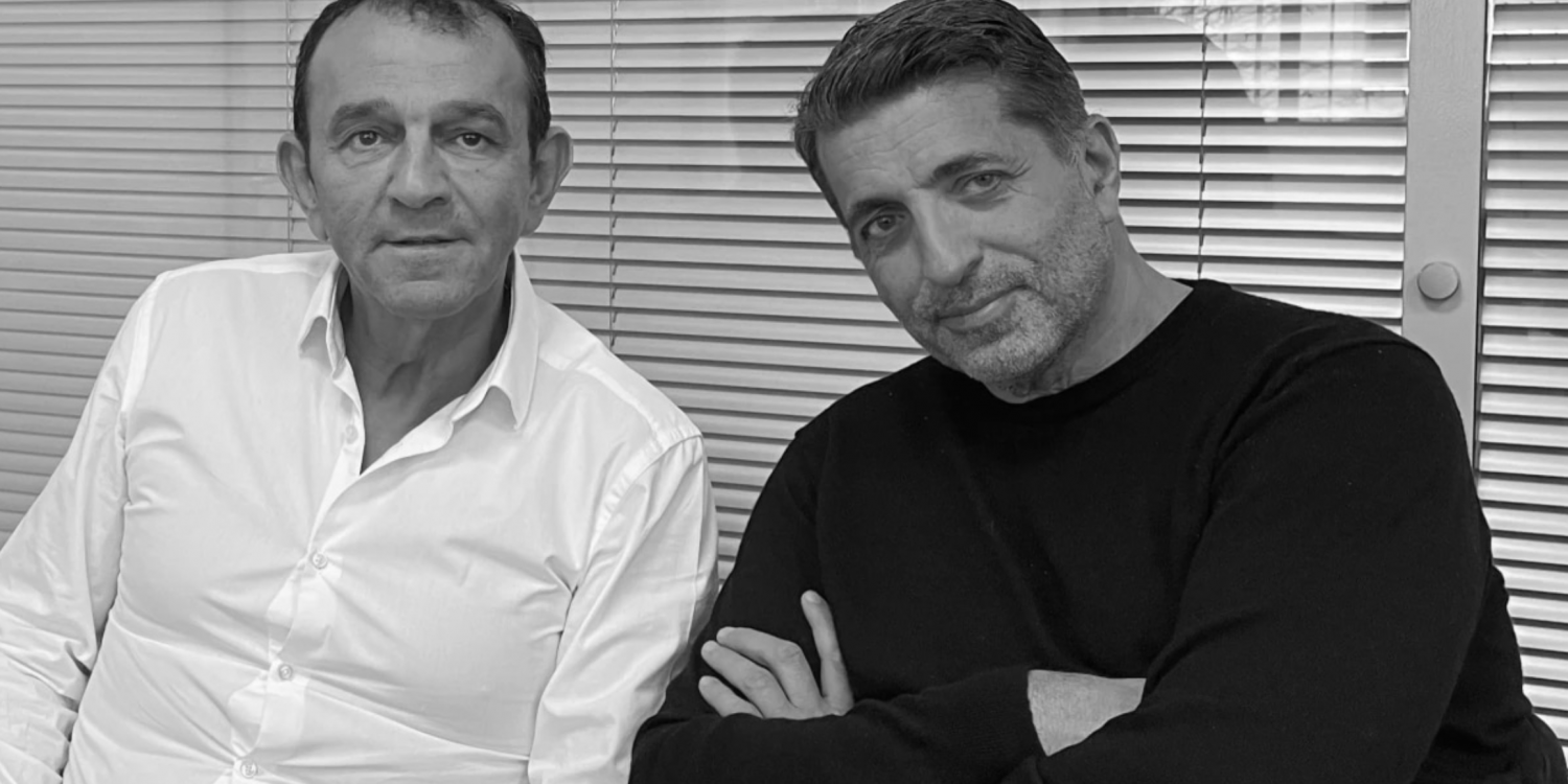 Two men sit side by side in front of window blinds; left man in a white shirt, right man in a black sweater, looking at the camera.