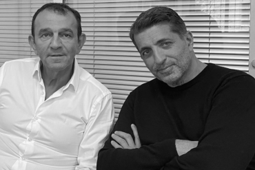 Two men sit side by side in front of window blinds; left man in a white shirt, right man in a black sweater, looking at the camera.