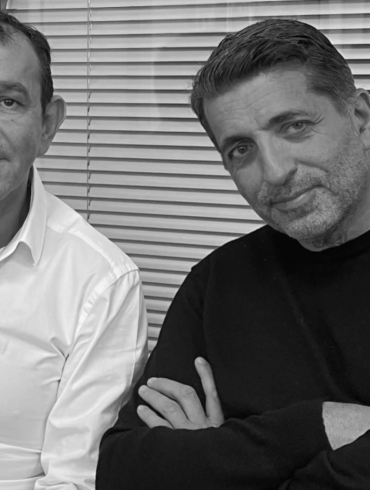 Two men sit side by side in front of window blinds; left man in a white shirt, right man in a black sweater, looking at the camera.