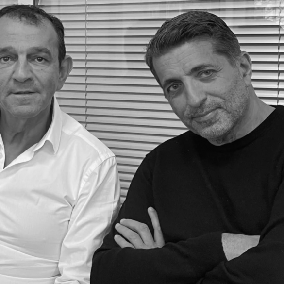 Two men sit side by side in front of window blinds; left man in a white shirt, right man in a black sweater, looking at the camera.
