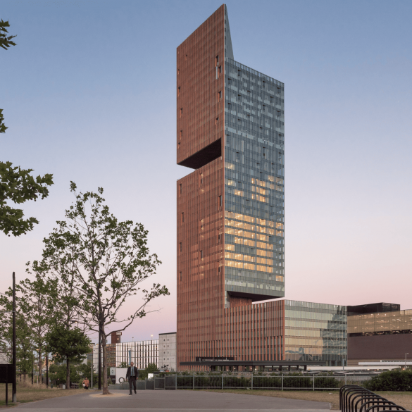 Tall modern tower with stacked brick and glass sections at sunset; trees in the foreground and a person walking nearby.