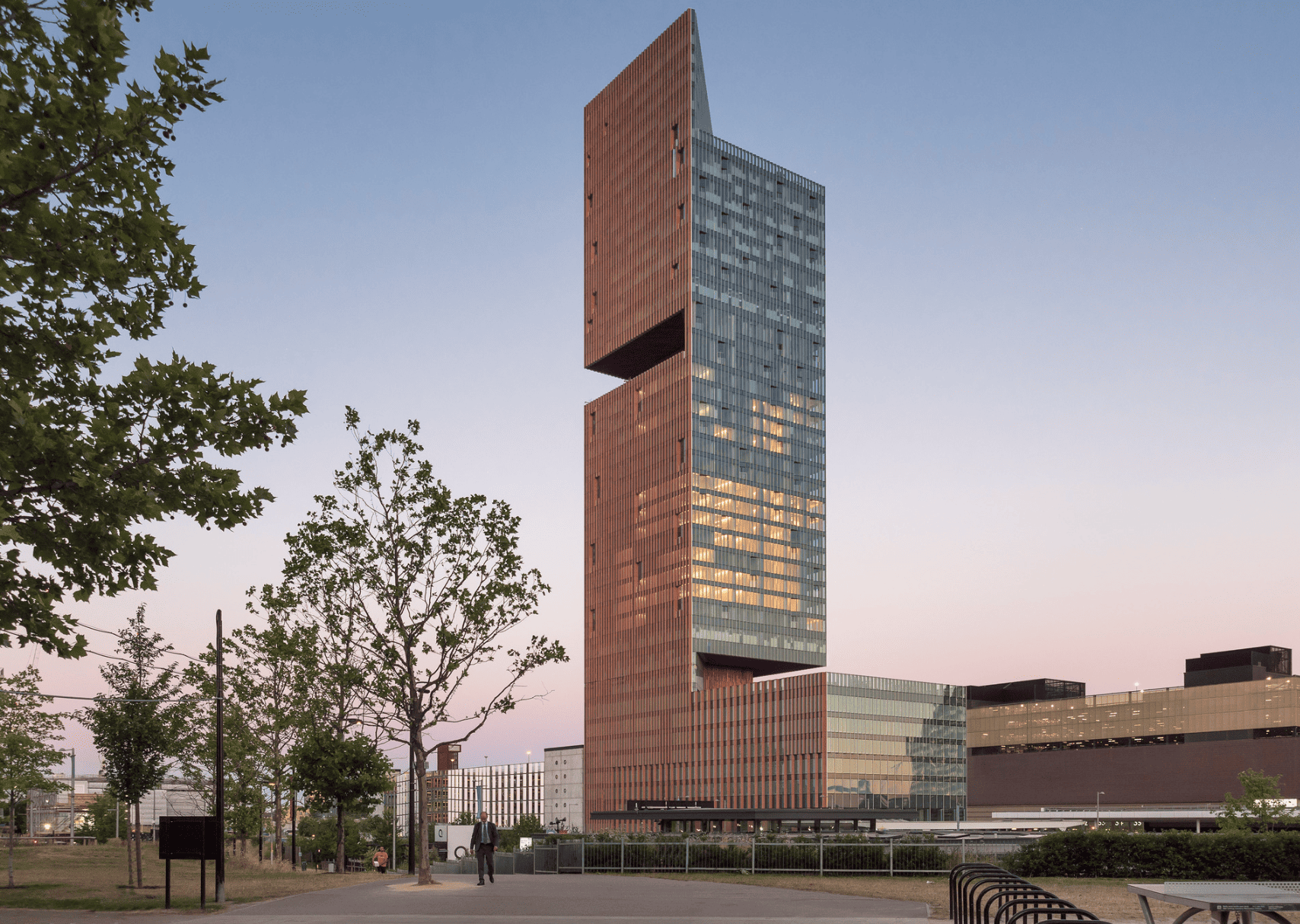 Tall modern tower with stacked brick and glass sections at sunset; trees in the foreground and a person walking nearby.