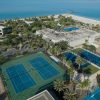 Aerial view of a tropical resort with a large pool area, palm trees, tennis courts, and a sandy beach along bright blue water.