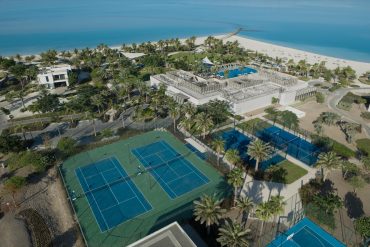 Aerial view of a tropical resort with a large pool area, palm trees, tennis courts, and a sandy beach along bright blue water.