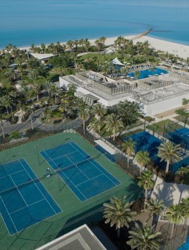 Aerial view of a tropical resort with a large pool area, palm trees, tennis courts, and a sandy beach along bright blue water.