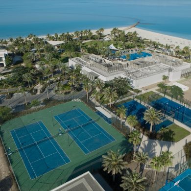 Aerial view of a tropical resort with a large pool area, palm trees, tennis courts, and a sandy beach along bright blue water.