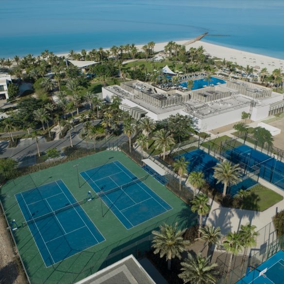 Aerial view of a tropical resort with a large pool area, palm trees, tennis courts, and a sandy beach along bright blue water.
