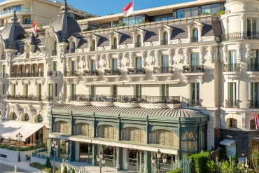 Ornate European-style luxury hotel on a palm-lined seaside street with iron balconies and intricate stonework.
