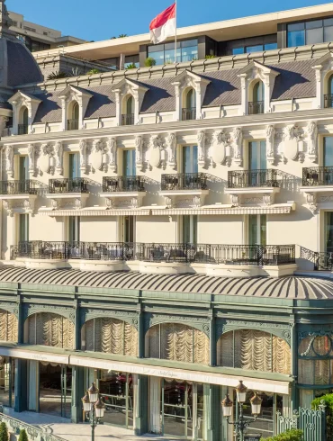 Ornate European-style luxury hotel on a palm-lined seaside street with iron balconies and intricate stonework.