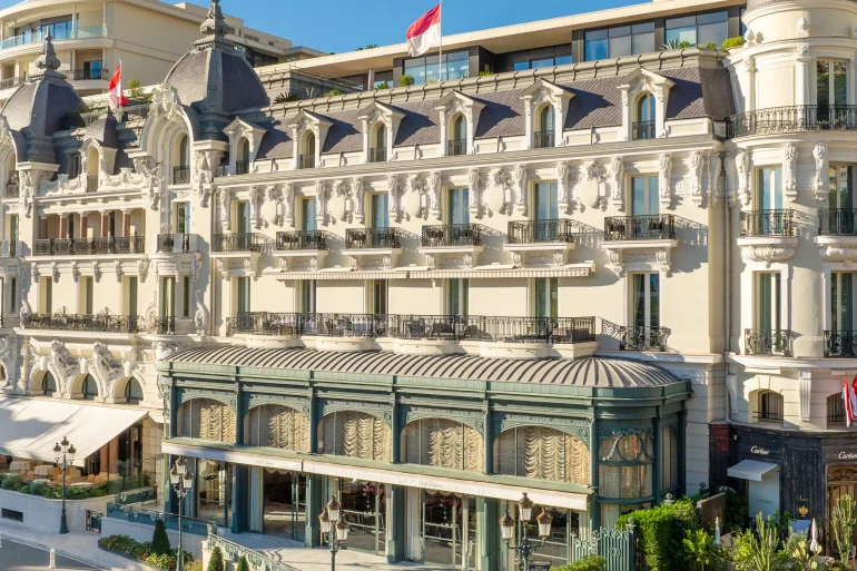 Ornate European-style luxury hotel on a palm-lined seaside street with iron balconies and intricate stonework.