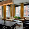 Bright living room with large windows, mustard-yellow curtains, beige sectional sofa, and a marble coffee table overlooking city brick buildings.