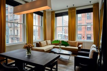 Bright living room with large windows, mustard-yellow curtains, beige sectional sofa, and a marble coffee table overlooking city brick buildings.