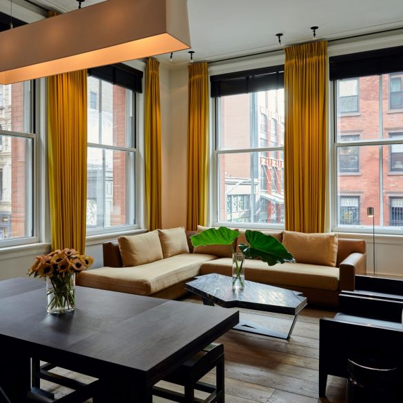 Bright living room with large windows, mustard-yellow curtains, beige sectional sofa, and a marble coffee table overlooking city brick buildings.