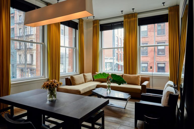 Bright living room with large windows, mustard-yellow curtains, beige sectional sofa, and a marble coffee table overlooking city brick buildings.