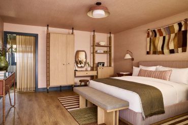 Hotel-style bedroom with a large bed, wooden bench at the foot, beige headboard, and midcentury furniture in warm earth tones.