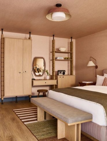 Hotel-style bedroom with a large bed, wooden bench at the foot, beige headboard, and midcentury furniture in warm earth tones.