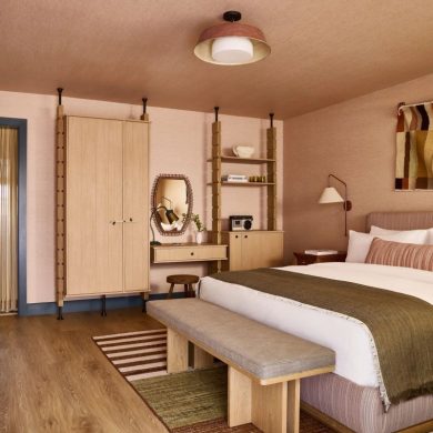 Hotel-style bedroom with a large bed, wooden bench at the foot, beige headboard, and midcentury furniture in warm earth tones.