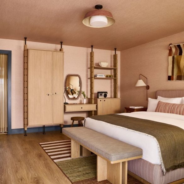 Hotel-style bedroom with a large bed, wooden bench at the foot, beige headboard, and midcentury furniture in warm earth tones.