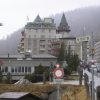 Urban street scene in a mountain town: a large stone hotel with green, pointed roofs, a construction crane on the left, and pedestrians nearby.