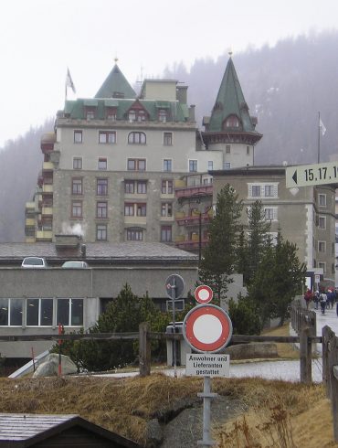Urban street scene in a mountain town: a large stone hotel with green, pointed roofs, a construction crane on the left, and pedestrians nearby.