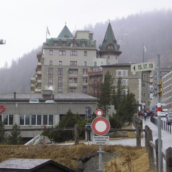 Urban street scene in a mountain town: a large stone hotel with green, pointed roofs, a construction crane on the left, and pedestrians nearby.