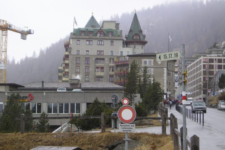 Urban street scene in a mountain town: a large stone hotel with green, pointed roofs, a construction crane on the left, and pedestrians nearby.