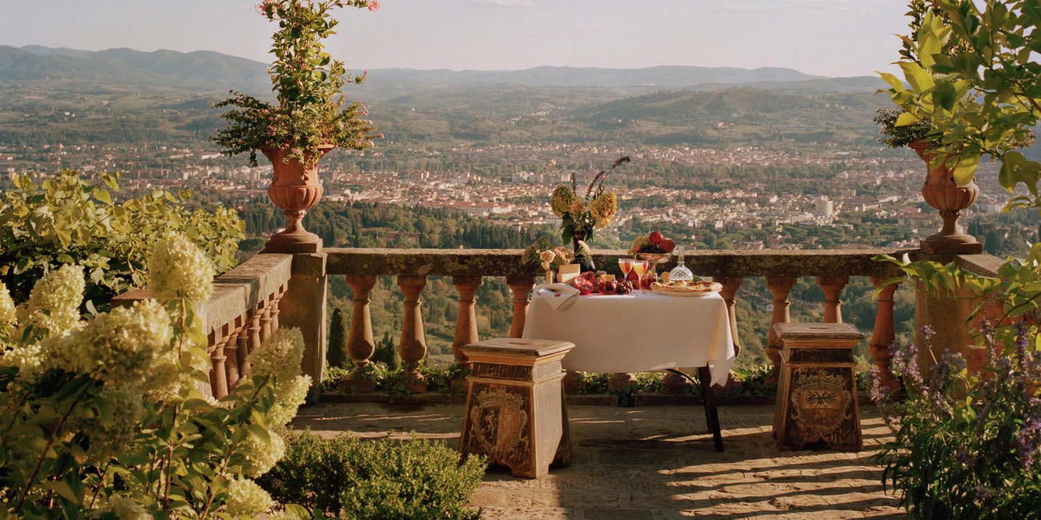 Table set for a meal on a stone terrace with potted plants and a view of a valley and distant hills