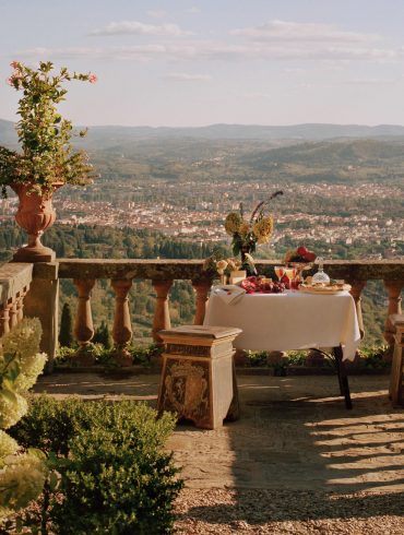 Table set for a meal on a stone terrace with potted plants and a view of a valley and distant hills