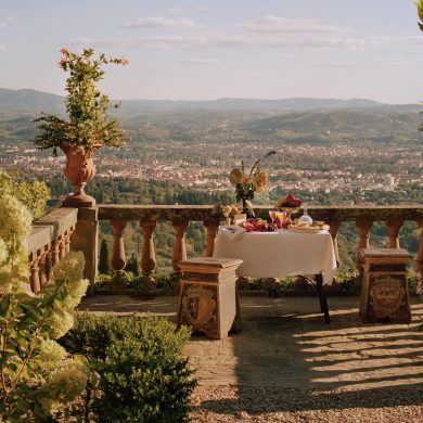 Table set for a meal on a stone terrace with potted plants and a view of a valley and distant hills