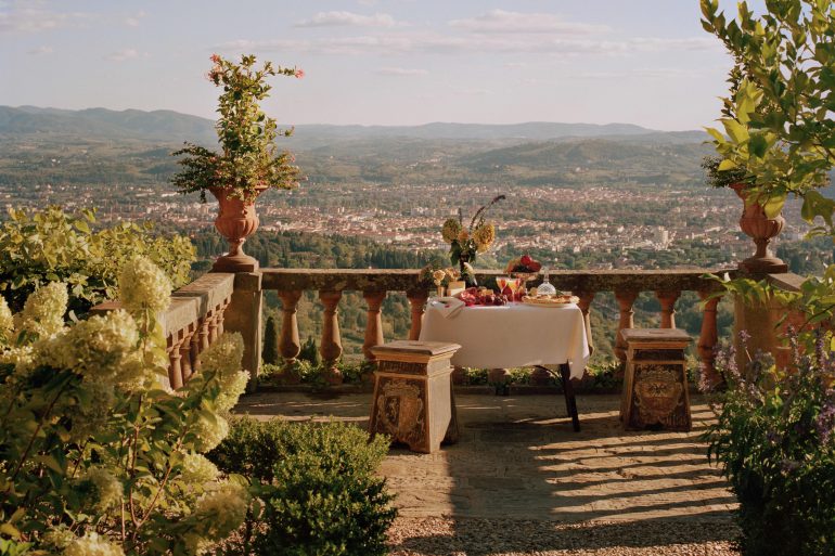 Table set for a meal on a stone terrace with potted plants and a view of a valley and distant hills