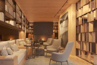 Cozy modern library lounge with built‑in wooden shelves, warm lighting, and a central fireplace flanked by beige seating.