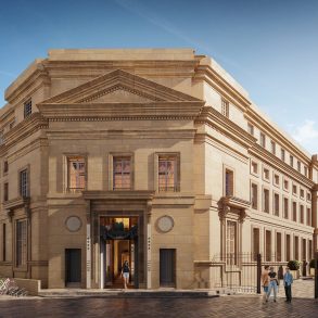 Beige stone hotel with a grand, ornate facade and a prominent triangular pediment, people standing and walking outside on a sunny day.