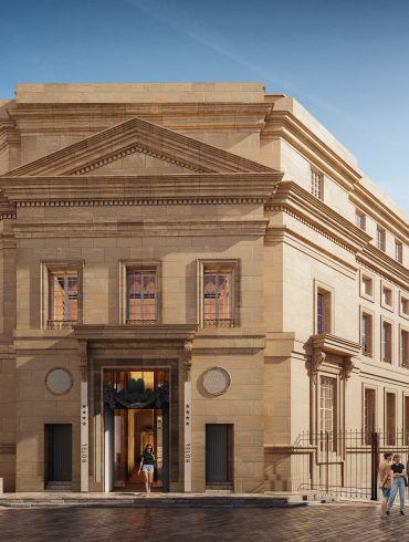 Beige stone hotel with a grand, ornate facade and a prominent triangular pediment, people standing and walking outside on a sunny day.