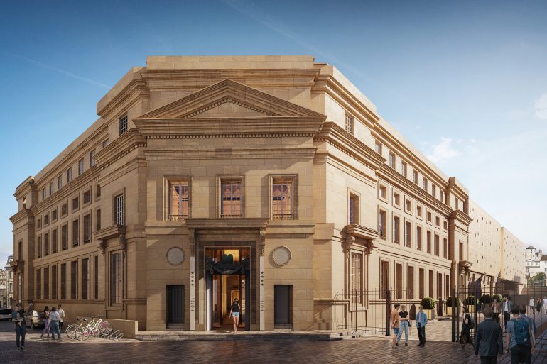 Beige stone hotel with a grand, ornate facade and a prominent triangular pediment, people standing and walking outside on a sunny day.