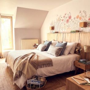 Cozy bedroom with a white duvet, beige throw, and decorative pillows on a wooden bed beneath a pastel floral mural above the headboard.
