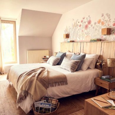 Cozy bedroom with a white duvet, beige throw, and decorative pillows on a wooden bed beneath a pastel floral mural above the headboard.