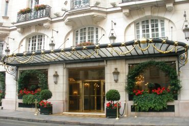 Hotel entrance with ornate stone facade, gold-accented canopy, and red flowering planters outside Le Bristol Paris (pinkish beige building).