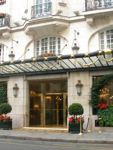 Hotel entrance with ornate stone facade, gold-accented canopy, and red flowering planters outside Le Bristol Paris (pinkish beige building).