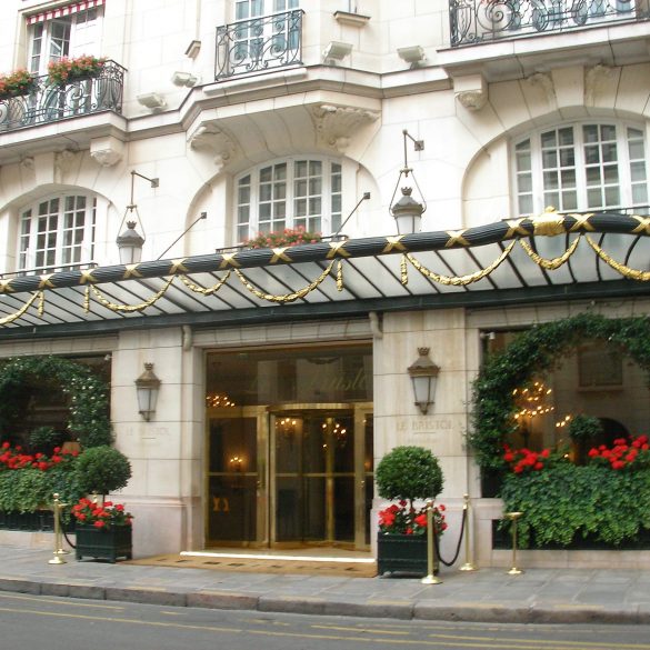Hotel entrance with ornate stone facade, gold-accented canopy, and red flowering planters outside Le Bristol Paris (pinkish beige building).
