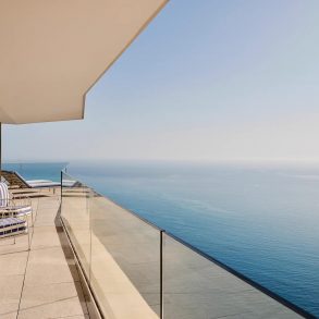 Modern balcony overlooking the ocean with glass railing and blue‑striped lounge chairs along a tiled terrace.