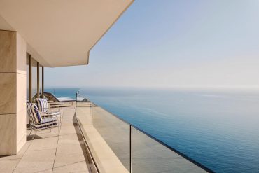 Modern balcony overlooking the ocean with glass railing and blue‑striped lounge chairs along a tiled terrace.