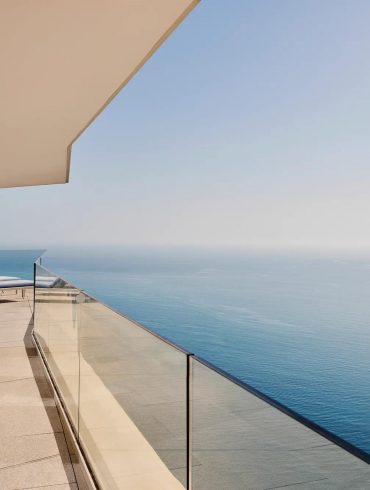 Modern balcony overlooking the ocean with glass railing and blue‑striped lounge chairs along a tiled terrace.